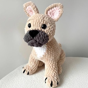 Könnte beinhalten: Ein handgefertigter, plüschiger Französische Bulldogge Hund. Der Hund ist beige mit einer grauen Schnauze und schwarzen Augen und Pfoten. Die Innenseite der Ohren ist rosa. Der Hund sitzt auf einer weißen Oberfläche.