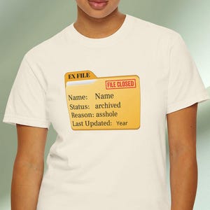 Puede incluir: Camiseta color crema con un diseño gráfico de una carpeta amarilla con detalles rojos y el texto "FILE CLOSED". La carpeta muestra "EX FILE" y contiene el texto "Name: Name, Status: archived, Reason: asshole, Last Updated: Year."