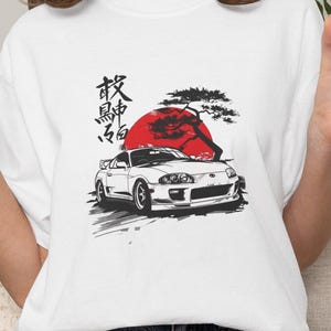 Könnte beinhalten: Weißes T-Shirt mit einem Aufdruck eines weißen Sportwagens, einer roten Sonne und japanischem Text. Das Design ist in Schwarz und Rot gehalten. Das T-Shirt wird mit Jeansshorts getragen.