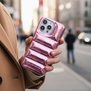 Op de afbeelding: Een roze telefoonhoesje met een gewatteerd ontwerp wordt in een hand gehouden. De hoes heeft een glanzende afwerking en de tekst "VEGAN SOPHISTICATION". Het heeft drie cameralenzen en een flitser.