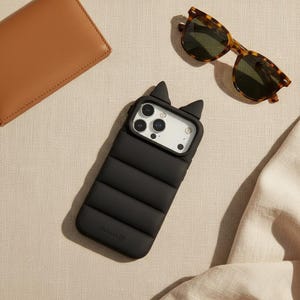 May include: Black iPhone case with cat ear design, featuring a quilted pattern. Tortoiseshell sunglasses and a brown leather wallet are also visible. The phone case has the brand name "VOLKERK" printed on it.