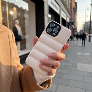 Peut inclure: Un étui de téléphone beige matelassé avec un module caméra est tenu dans une main. L'étui présente un motif à rayures horizontales rembourrées. L'étui de téléphone est de couleur neutre, et l'arrière-plan est une rue de la ville.
