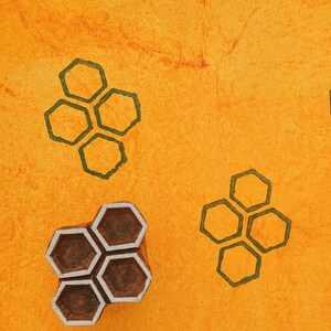 Puede incluir: Un fondo naranja con cuatro grupos de formas de panal verdes. Un sello de madera con un patrón de panal es visible en la esquina inferior izquierda. La imagen tiene una apariencia texturizada.