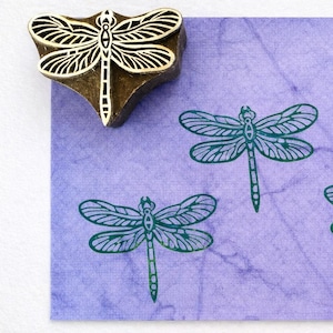 May include: A wooden dragonfly stamp next to three teal dragonfly impressions on a purple textured paper. The stamp is a dark brown color with a dragonfly design carved into the bottom. The paper has a marbled effect.
