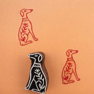 Op de afbeelding: Een houten hondenstempel met een zwart-wit ontwerp, naast verschillende rode hondenprints op een oranje achtergrond. De hondenprints hebben een bloemenpatroon op hun lichaam.