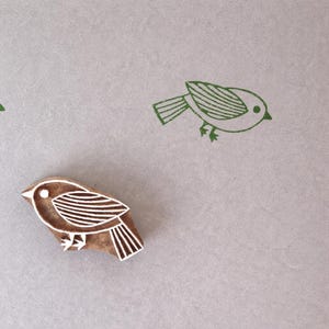 May include: A wooden bird-shaped printing block with carved line details on the wing and tail. Two green ink impressions of the same bird design are stamped above the wooden block, showing the bird facing toward the right side.