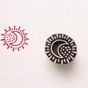 May include: A wooden stamp featuring a sun and crescent moon motif. The stamp is dark brown with white detailing. Two red ink impressions of the design flank the stamp, creating a celestial theme.