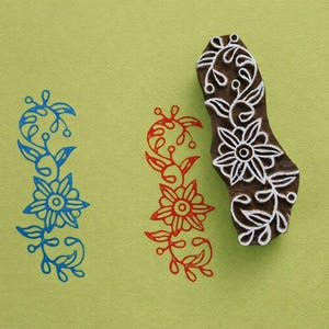 Könnte beinhalten: Ein Holzstempel mit einem Blumendesign in weißer Tinte. Der Stempel befindet sich neben drei Blumenmustern in blauer und roter Tinte auf einem hellgrünen Hintergrund. Die Blumenmuster sind vertikal und zeigen Blumen und Blätter.
