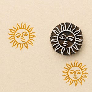 May include: A wooden sun stamp with a detailed sun face design is surrounded by three stamped sun images. The stamped images are in a golden yellow color, and the stamp itself is dark brown with white accents.