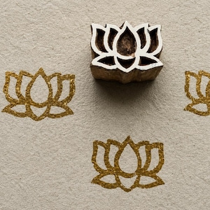 May include: A wooden block stamp featuring a carved lotus flower design, positioned above three identical lotus flower impressions stamped in a shimmering gold ink on a textured, light beige paper surface.