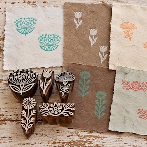 Puede incluir: Cinco sellos de madera tallada con intrincados patrones florales dispuestos junto a trozos de papel texturizado hecho a mano con diseños estampados en tinta turquesa, blanca, naranja y roja, mostrando varios motivos botánicos y formas florales.