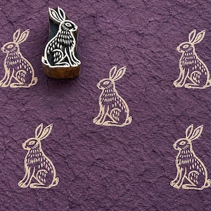 Op de afbeelding: Een houten stempel met een uitgesneden konijnontwerp, geplaatst boven vijf afdrukken van hetzelfde konijn in een lichtbeige kleur op een gestructureerd, donkerpaars papieroppervlak. De stempel toont een gedetailleerde, gestileerde haas.