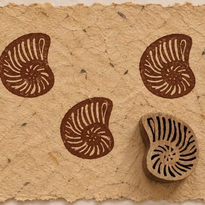 May include: A rectangular sheet of textured, light tan paper featuring two dark brown stamped nautilus shell designs and one wooden nautilus shell printing block positioned in the bottom right corner, showing the carved spiral pattern.