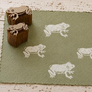 Puede incluir: Una hoja de papel rectangular de color verde oliva con cuatro ilustraciones de ranas impresas en blanco. Dos sellos de madera marrón con tallas de rana a juego están colocados en la esquina superior izquierda del papel.