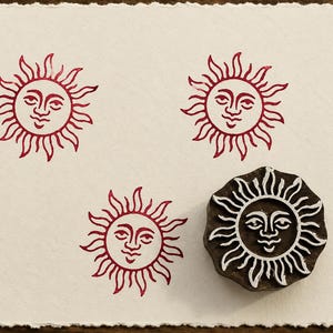 May include: A dark brown wooden sun-shaped printing block with a carved face and rays, positioned next to three red ink impressions of the same sun design stamped onto a square piece of off-white paper with a deckled edge.