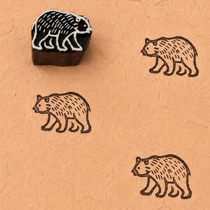 May include: A dark brown wooden block stamp featuring a carved bear design, positioned above five identical brown ink impressions of the same bear on a light tan textured paper surface. The stamp itself shows the bear in white relief.