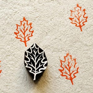 May include: A black wooden block stamp carved in the shape of an oak leaf, positioned centrally on a textured off-white paper surface. Surrounding the stamp are four orange ink impressions of the same leaf design, creating a repeating pattern.