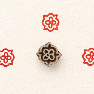 Può includere: Un timbro in legno con un motivo floreale, circondato da tre impressioni floreali rosse. Il timbro è marrone con un motivo dettagliato e le impressioni sono di un rosso brillante. Lo sfondo è una superficie bianco sporco strutturata.