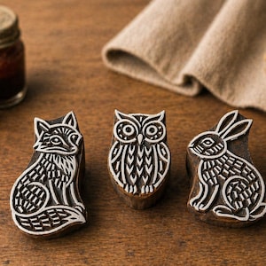 May include: Three wooden animal stamps: a fox, an owl, and a rabbit. Each stamp is carved with intricate white designs on a brown background. The fox is seated, the owl is centered, and the rabbit is seated. Small jars and a tan cloth are in the background.