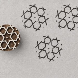 Puede incluir: Un sello de madera circular con un patrón geométrico en relieve de contornos hexagonales. El sello se encuentra junto a tres impresiones en tinta negra del mismo diseño, que consiste en un punto central rodeado por seis hexágonos.