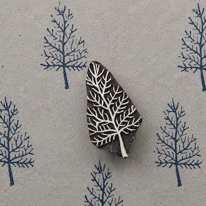 May include: A dark brown wooden printing block carved with a detailed tree branch design, positioned over a light grey surface featuring a repeating pattern of dark blue printed trees that match the shape of the carved wooden block.