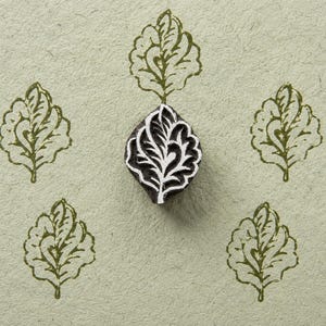 May include: A dark wooden block stamp with a carved leaf pattern positioned in the center, surrounded by four identical olive green leaf prints stamped onto a textured light beige paper surface, showcasing the intricate details of the carved design.