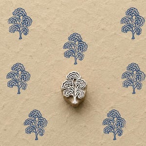 May include: A wooden printing block carved in the shape of a tree with a rounded canopy, positioned centrally on a textured tan surface surrounded by seven identical blue ink impressions of the same tree design arranged in a symmetrical pattern.