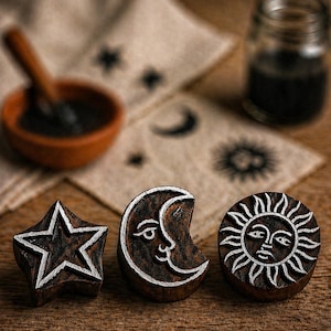 May include: Three wooden stamps featuring celestial designs: a star, a crescent moon with a face, and a sun with a face. Each stamp has a dark brown base with white detailing. The stamps are on a wooden surface, with ink and a bowl in the background.