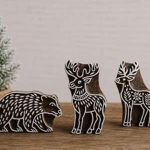 May include: Three dark brown wooden printing blocks featuring carved animal designs. One block depicts a bear in profile, while the other two blocks show standing deer with intricate patterns on their bodies. The carvings have white outlines.