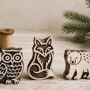 Könnte beinhalten: Drei Holzstempel mit Tieren: eine Eule, ein Fuchs und ein Bär. Die Stempel sind braun mit weißen Details und auf einer hellen Holzoberfläche angeordnet. Die Eule hat große Augen, der Fuchs sitzt und der Bär steht.
