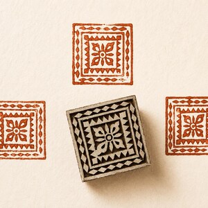 May include: A wooden block with a square design, surrounded by three stamped impressions in a reddish-brown hue. The block features a floral pattern within a geometric border, creating a decorative effect. The stamped impressions are also square with a similar design.