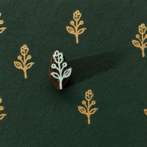 May include: A dark green surface with a repeating pattern of gold floral designs. A wooden stamp with a floral design in white and light blue is in the center. The stamp's shadow is visible.