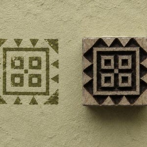 May include: A square wooden printing block featuring a geometric pattern of a central square divided into four smaller squares with a dot in the middle, surrounded by a border of triangles, shown with a matching olive green ink stamp impression.