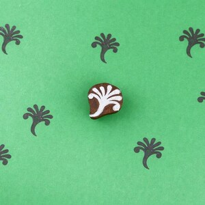 Puede incluir: Un bloque de madera marrón con un diseño floral blanco está centrado sobre un fondo verde. Ocho impresiones de sellos florales negros rodean el bloque. Adecuado para proyectos de manualidades e impresión.