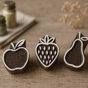 May include: Three wooden fruit-shaped stamps: an apple, a strawberry, and a pear. Each stamp is dark brown with a white outline and details. The apple and pear have leaves, while the strawberry features a textured pattern. The stamps are on a wooden surface.