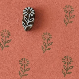 Op de afbeelding: Een houten stempel met een uitgesneden madeliefje in een witte en donkerbruine afwerking, geplaatst op een getextureerd terracotta-kleurig oppervlak met meerdere gestempelde afdrukken van dezelfde bloem in een gedempt olijfgroen.