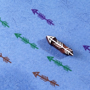 Puede incluir: Un sello de flecha de madera con un diseño de flecha blanca, rodeado de flechas estampadas en verde, marrón y morado sobre una superficie azul texturizada. Suministros para manualidades.