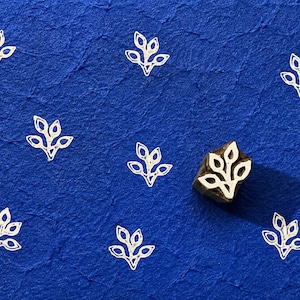 Puede incluir: Un sello de bloque de madera con un diseño de hoja blanca se encuentra sobre una superficie azul texturizada, junto a varios patrones de hojas estampadas. La sombra del sello es visible. El patrón de hojas se repite.