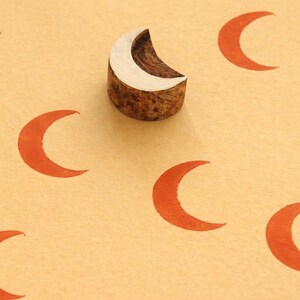 May include: A wooden crescent moon stamp with a natural bark edge, positioned among several stamped orange crescent moons on a light tan surface. The stamp is used for crafting and art projects.