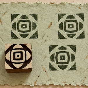 May include: A square wooden printing block featuring a geometric pattern of concentric squares and curved shapes, shown alongside three dark green ink impressions of the same design stamped onto a piece of light green textured handmade paper.