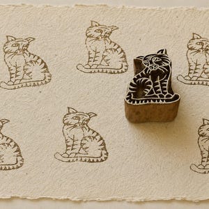 May include: A carved wooden block in the shape of a seated cat, positioned on a textured off-white paper surface. The block is shown next to several dark brown ink impressions of the same cat design, creating a repeating pattern of feline figures.