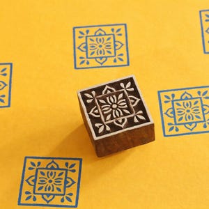 May include: A wooden block stamp with a floral design, surrounded by blue square patterns on a yellow background. The stamp is used to create the blue patterns. The design is a square within a square with a floral pattern.