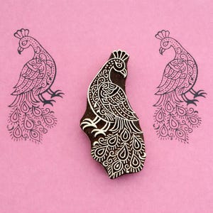 Puede incluir: Un bloque de madera con forma de pavo real con intrincados diseños blancos, flanqueado por dos contornos de pavo real sobre un fondo rosa. El bloque está tallado con patrones detallados, ideal para la impresión textil o proyectos de artesanía. El diseño del pavo real presenta una cola detallada y una corona.