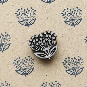 May include: A wooden block print stamp featuring a stylized flower design with a rounded head and two leaves. The stamp is positioned over a beige paper surface that displays a repeating pattern of the same blue flower motif printed in ink.