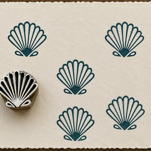 May include: A metal stamp shaped like a fan-style seashell with a silver finish, positioned next to a cream-colored paper featuring five dark teal ink impressions of the same seashell design arranged in a pattern across the surface.