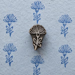 Op de afbeelding: Een donkerbruine houten blokstempel met een uitgesneden bloemmotief inclusief steel en bladeren. De stempel ligt op een lichtblauw papieren oppervlak dat verschillende blauwe inkt-afdrukken van hetzelfde bloempatroon in een raster toont.