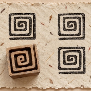 May include: A square wooden block stamp with a spiral pattern rests on a piece of textured, off-white handmade paper. The paper features three black ink impressions of the same square spiral design, each showing a consistent, repeating geometric motif.