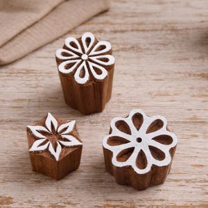 Pode incluir: Três carimbos de bloco de madeira com desenhos florais brancos. Os carimbos são castanhos com detalhes pintados de branco. Os carimbos estão dispostos sobre uma superfície de madeira clara, com um pano bege dobrado ao fundo.