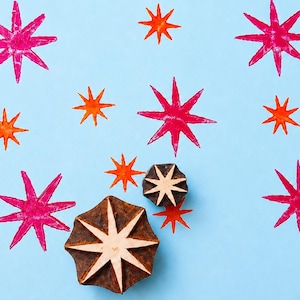 May include: A light blue background with hand-painted star designs in shades of pink and orange. Two wooden star-shaped stamps are also visible. The stamps have a brown exterior and a white star design.