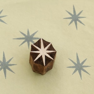Può includere: Un timbro in legno con un motivo a stella bianca e marrone, circondato da motivi a stella azzurra su sfondo crema. Lo strumento è utilizzato per creare motivi a stella ripetuti.
